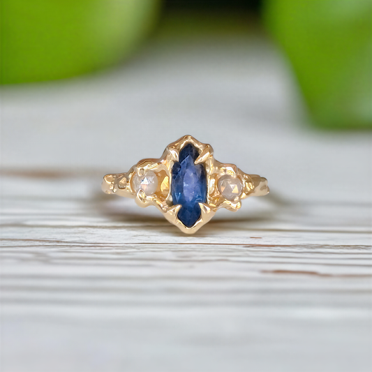 A gold ring featuring a central marquise-cut blue sapphire surrounded by small round diamonds, resting on a light wooden surface with a soft green background.