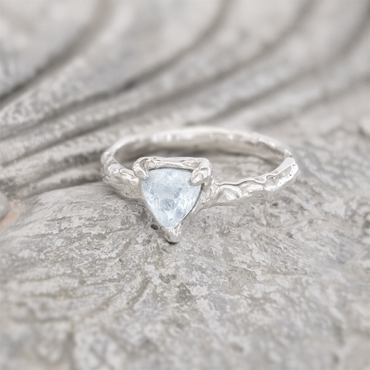 A silver ring with a blue gemstone, possibly a diamond, is displayed on a textured surface.