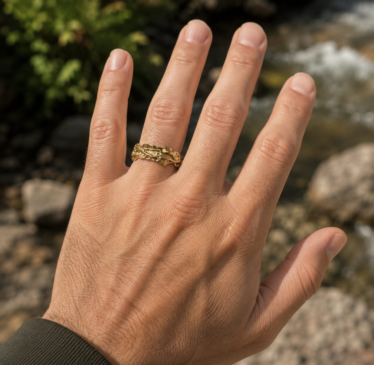 A close-up of a person's hand wearing a textured gold band ring outdoors near rocks and water.