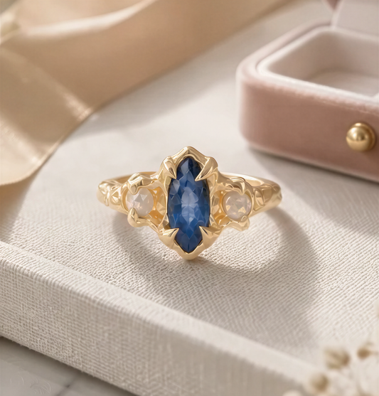 A gold ring with a blue gemstone, placed on a white surface, with a pink velvet box and some flowers in the background.