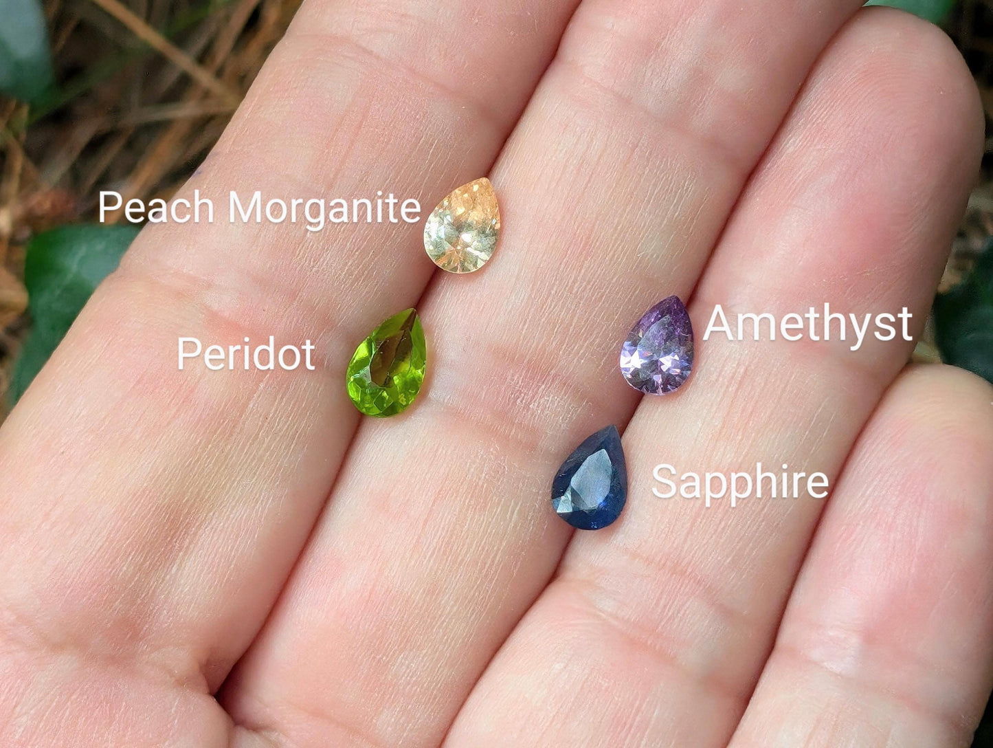 Hand holding gemstones: Peach Morganite, Peridot, Amethyst, and Sapphire in vibrant colors.