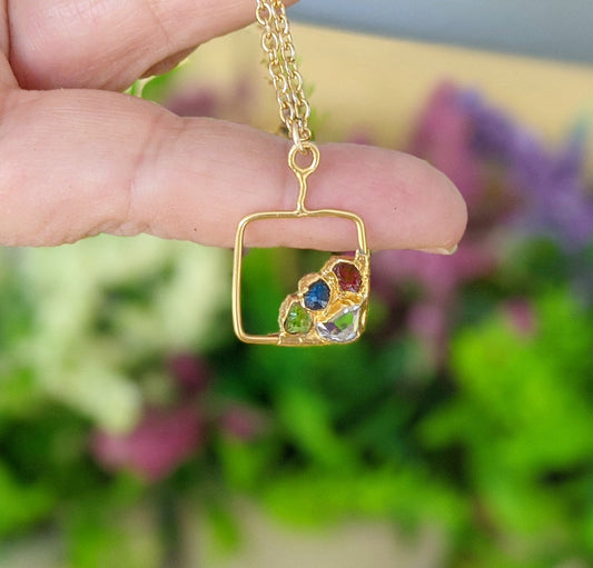 Gold-filled square pendant necklace with colorful raw gemstones, held against a blurred floral background.
