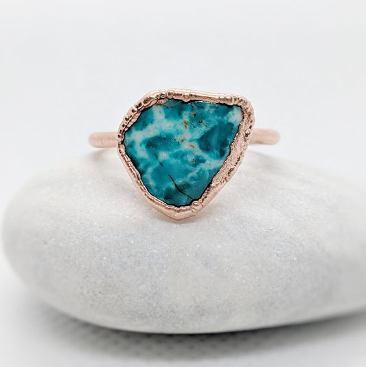 Natural turquoise ring with unique aqua-blue stone set in rose gold on a smooth white pebble.