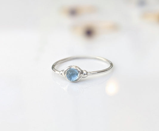 Dainty blue Topaz stacking ring on a reflective surface, featuring a 4mm natural light blue stone in a Sterling Silver band.
