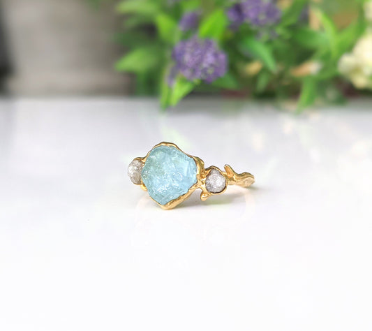 Raw Aquamarine and diamond twig ring on a white surface with green foliage in the background.
