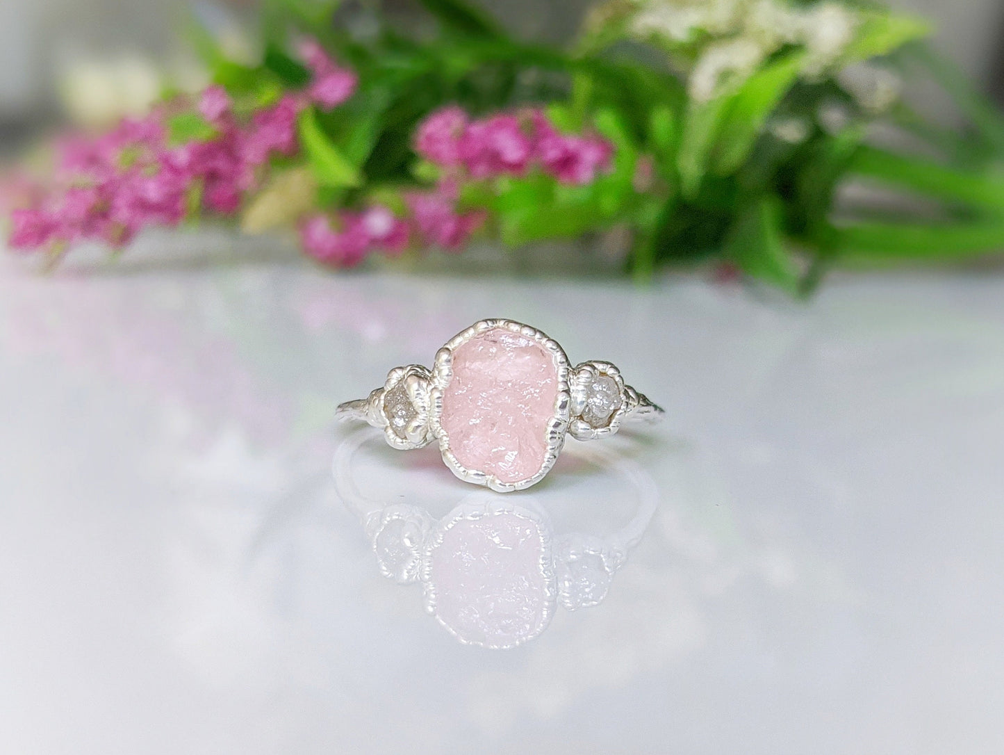 Raw Morganite ring with rough diamond and pink morganite on sterling silver band, set against floral background.