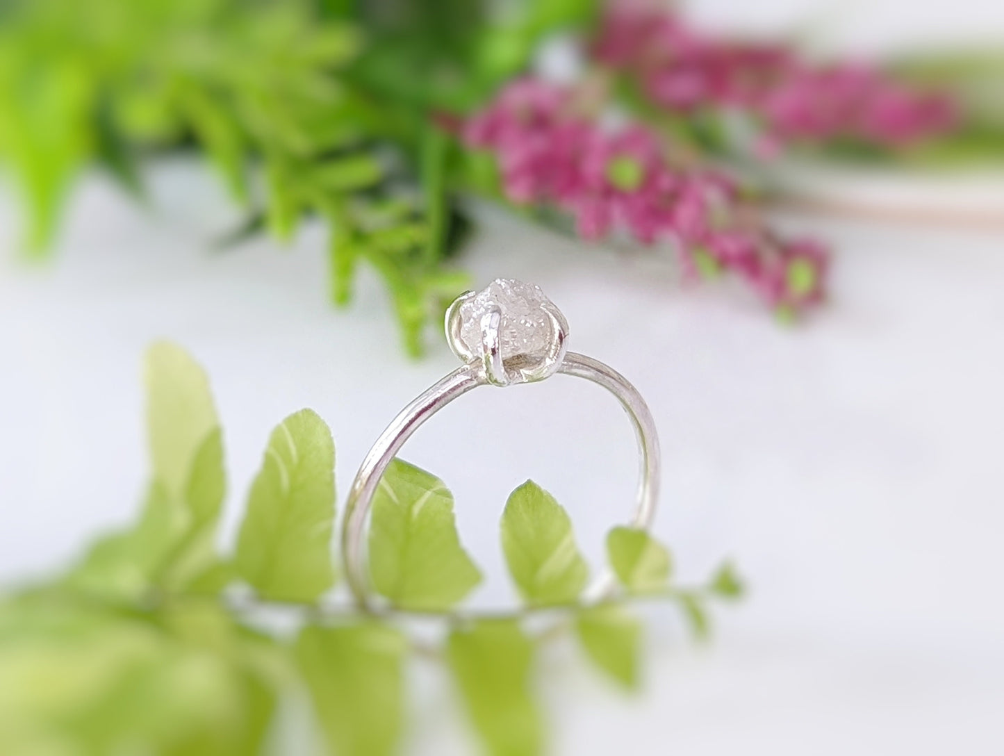 Elegant raw diamond engagement ring with a silver band, surrounded by greenery and soft floral elements.