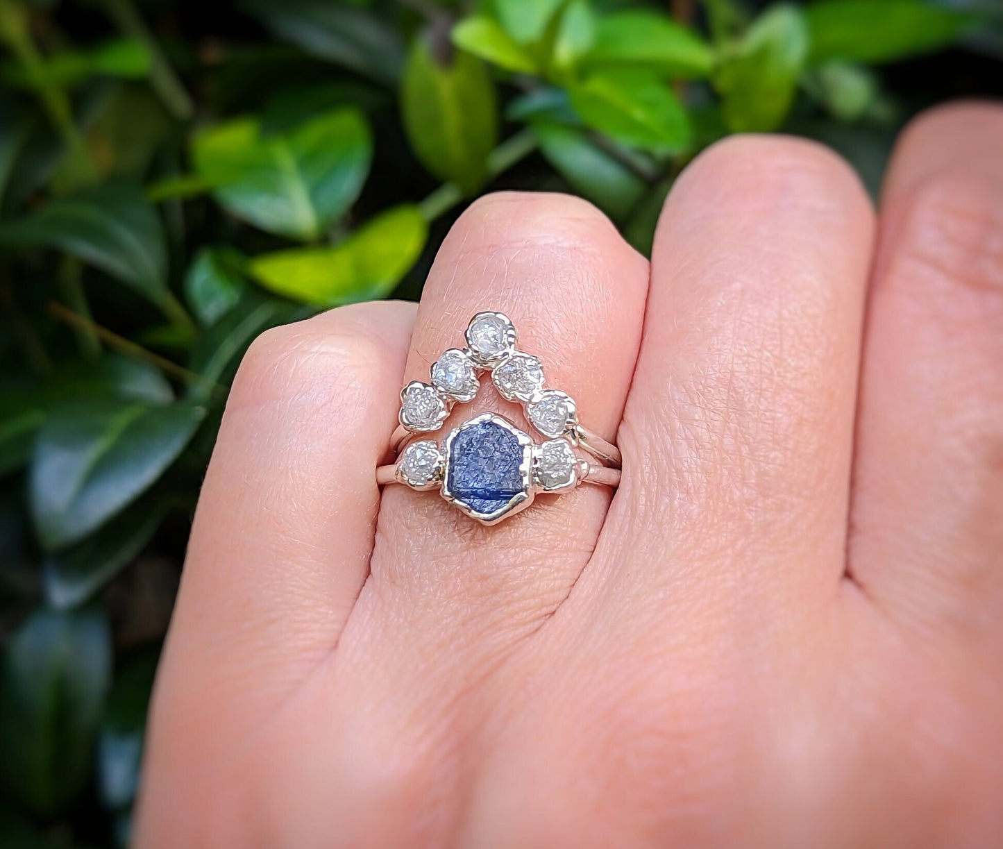 Raw sapphire and diamond wedding ring set showcased on a hand, surrounded by greenery.