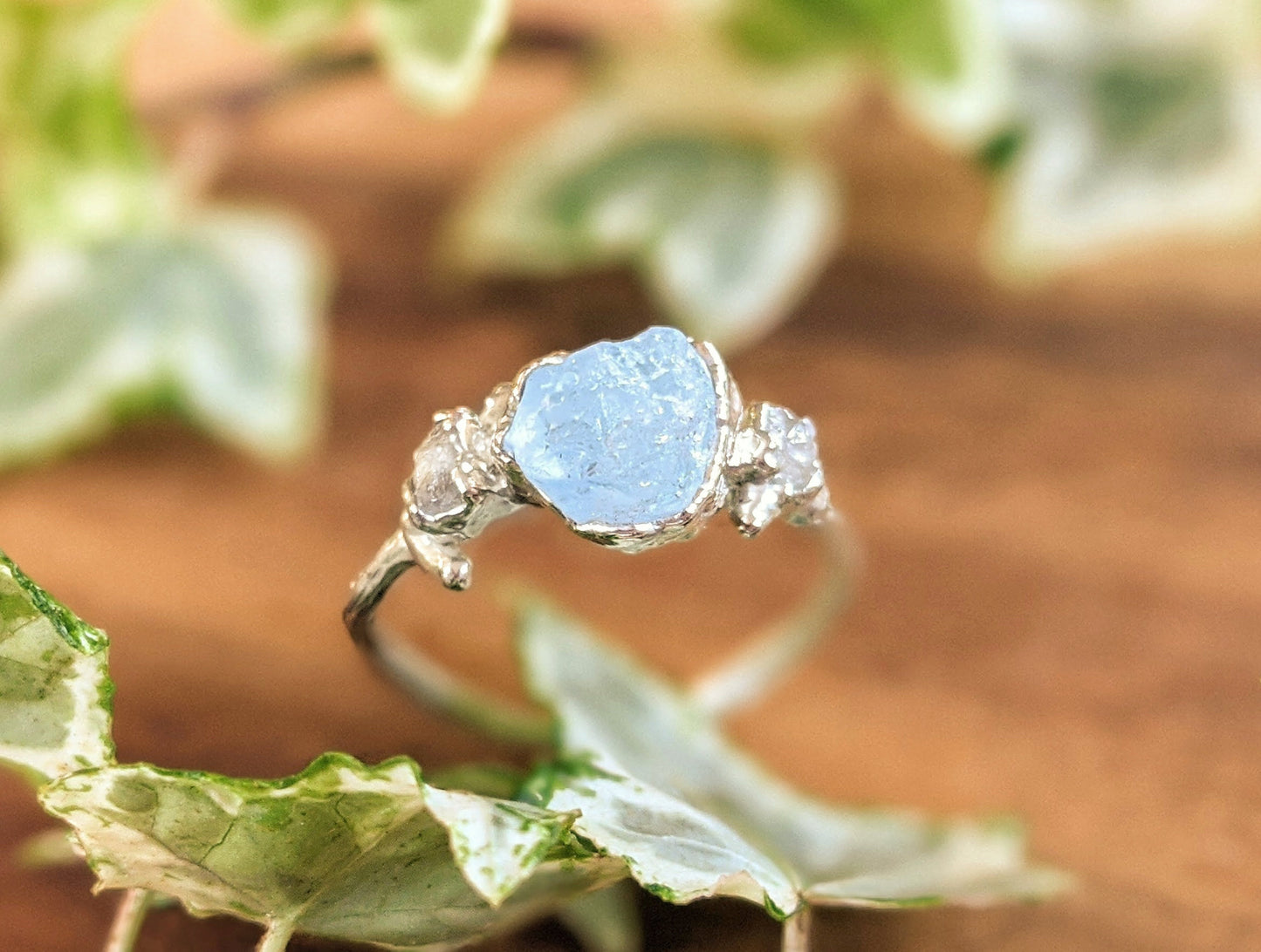 Raw aquamarine and diamond twig engagement ring on a wooden surface surrounded by green leaves.