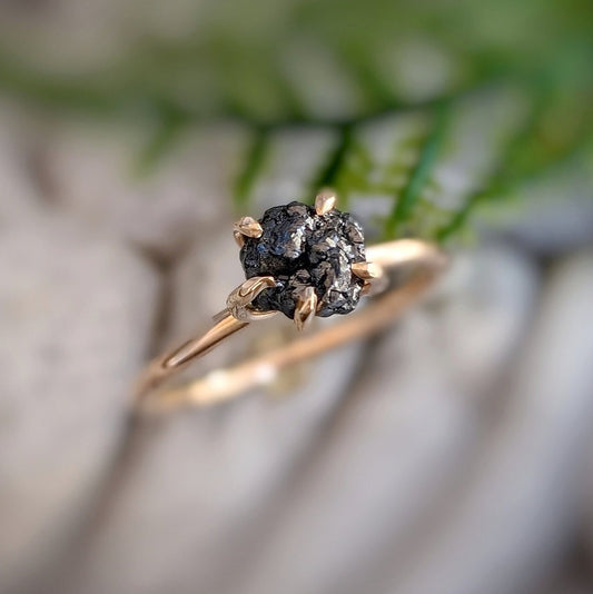 Raw black diamond engagement ring in solid 14k gold with a handcrafted prong band, showcased against a natural background.