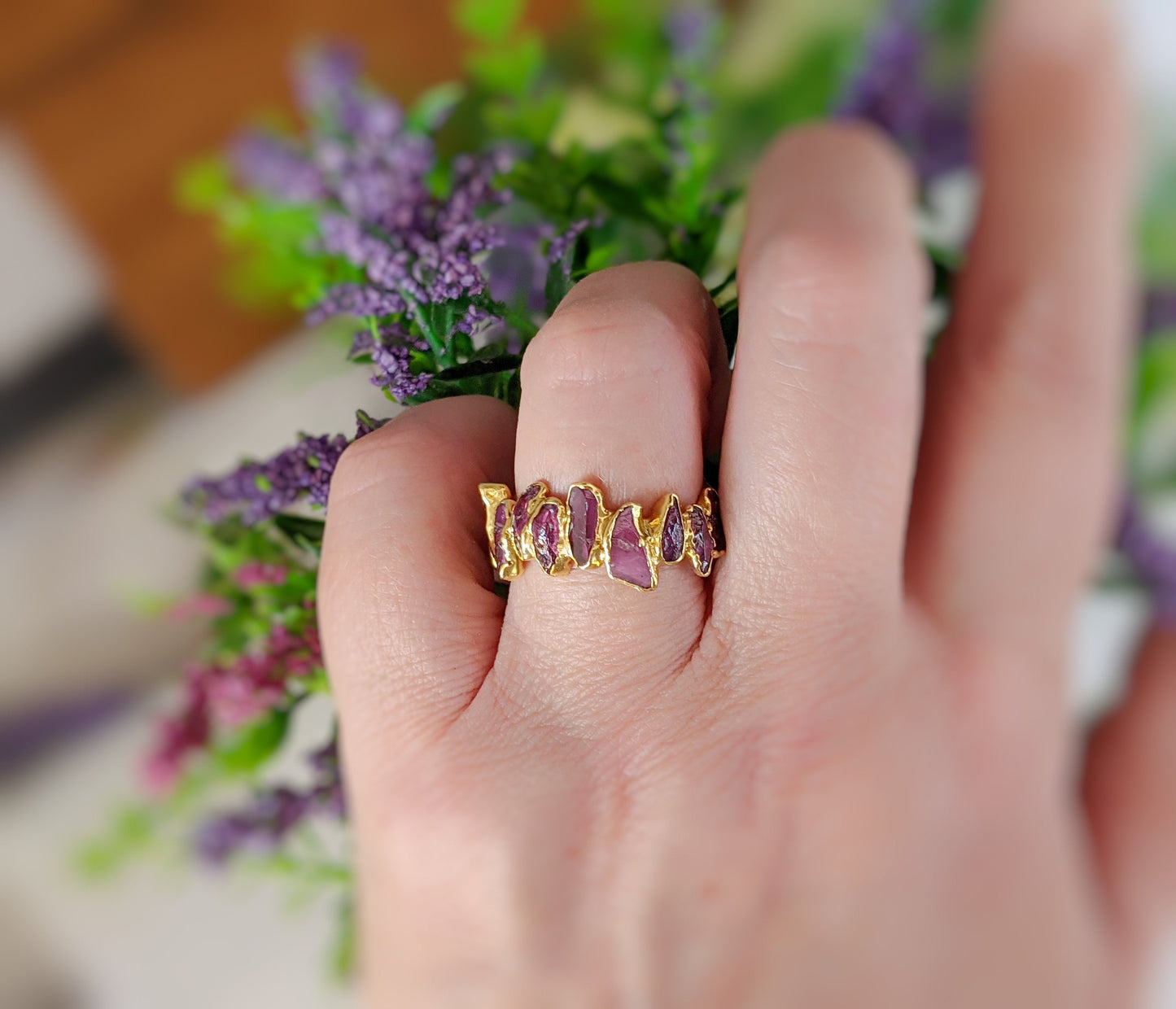 Raw Rhodolite Garnet Eternity Ring worn on a hand, showcasing its unique design and gemstones against a floral background.
