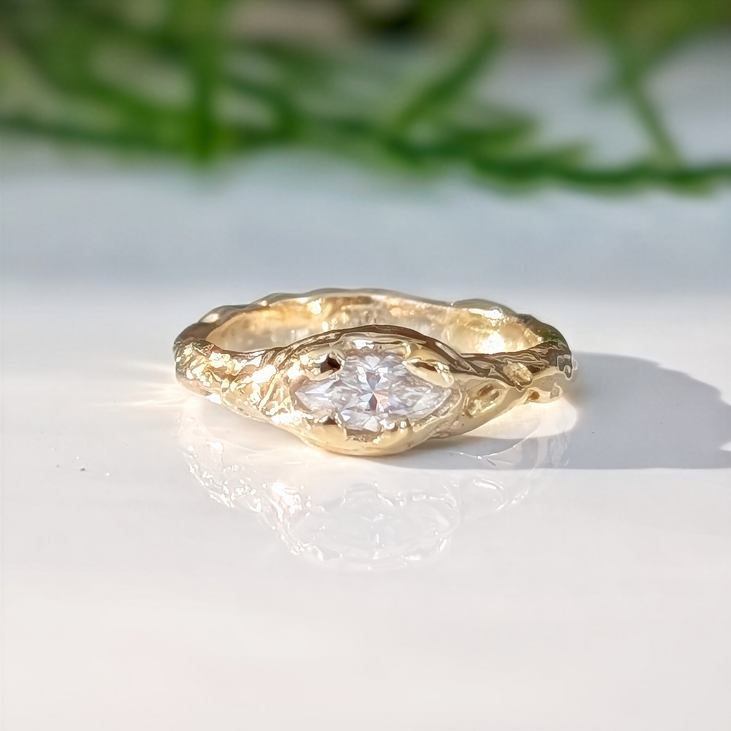 Gold ring with a diamond on a white surface with greenery in the background