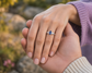 A silver ring with a marquise-cut blue Sapphire and small side diamonds, worn on a person's hand while another hand rests on top, set against a blurred outdoor background.