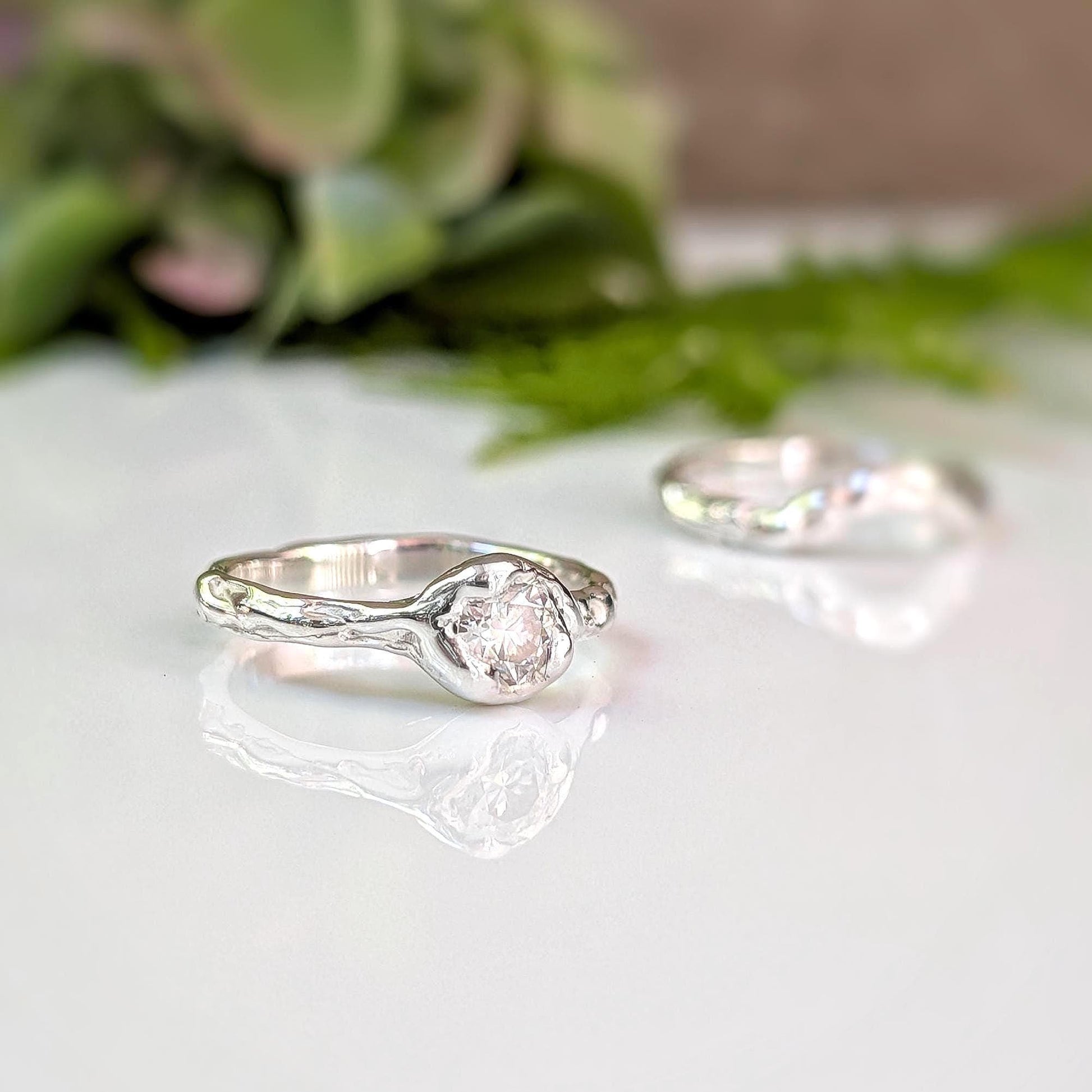 Two silver rings with a central stone are displayed on a white surface, with a blurred green plant in the background.