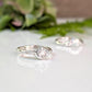 Two silver rings with a central stone are displayed on a white surface, with a blurred green plant in the background.