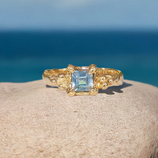 London Blue Topaz engagement ring in solid 14k gold with an organic textured band, set against a serene ocean backdrop.