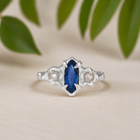A silver ring featuring a marquise-cut blue sapphire center stone and two small round clear stones on either side, set against a light beige background with soft green leaves in the upper background.