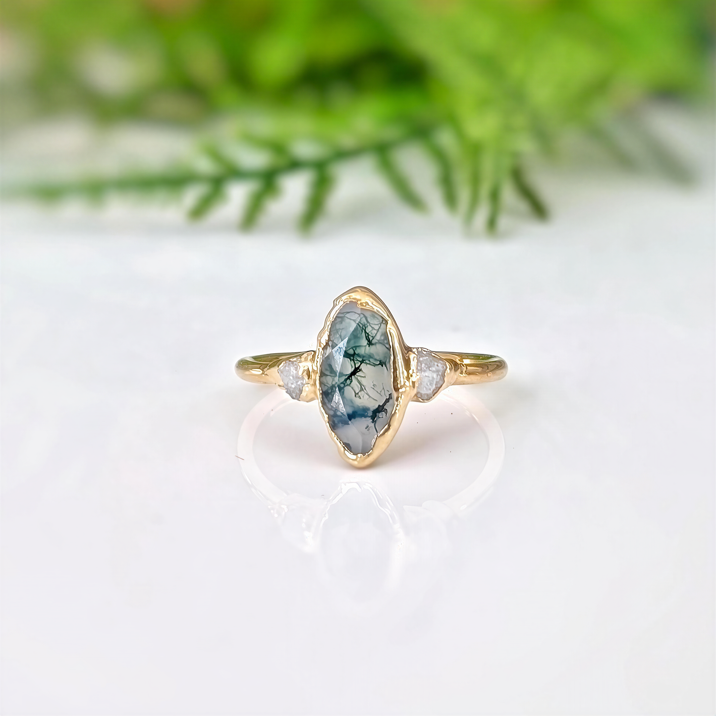Gold ring with a marquise shape Moss Agate and raw diamond engagement ring in a light background with some greenery