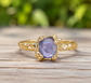 A gold ring with a large, round purple gemstone set in the center, placed on a wooden surface.