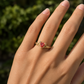 A textured gold ring with a bright orange sapphire worn on a person's finger, set against a blurred green outdoor background.