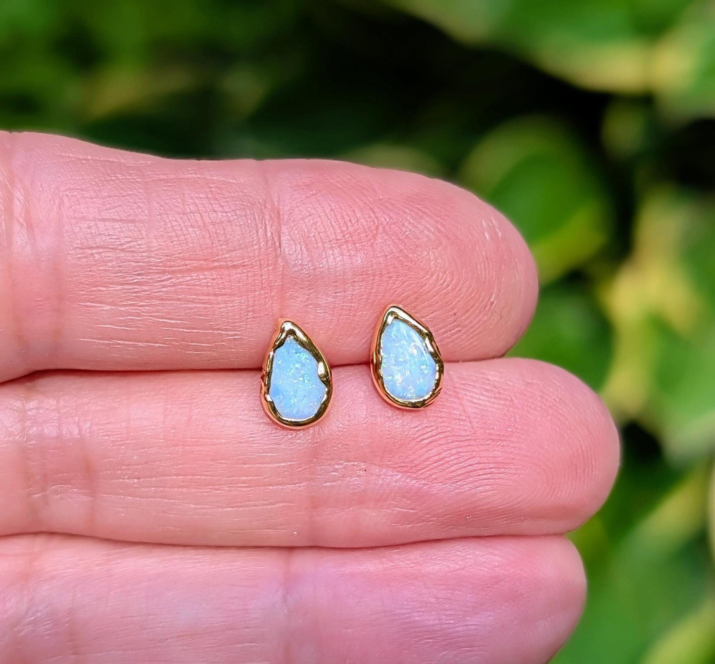 A hand holding a pair of pear-shape white australian opal stud earrings in gold textured setting. The opals have lovely flashes of color.