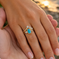 A gold engagement ring with a teardrop-shaped turquoise stone and small round diamonds, worn on a woman's hand which is held in a man's hand outdoors near the water at sunset.