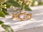 A gold ring with a large diamond set in the center, surrounded by a twisted, organic design, sits on a wooden surface with green leaves in the background.