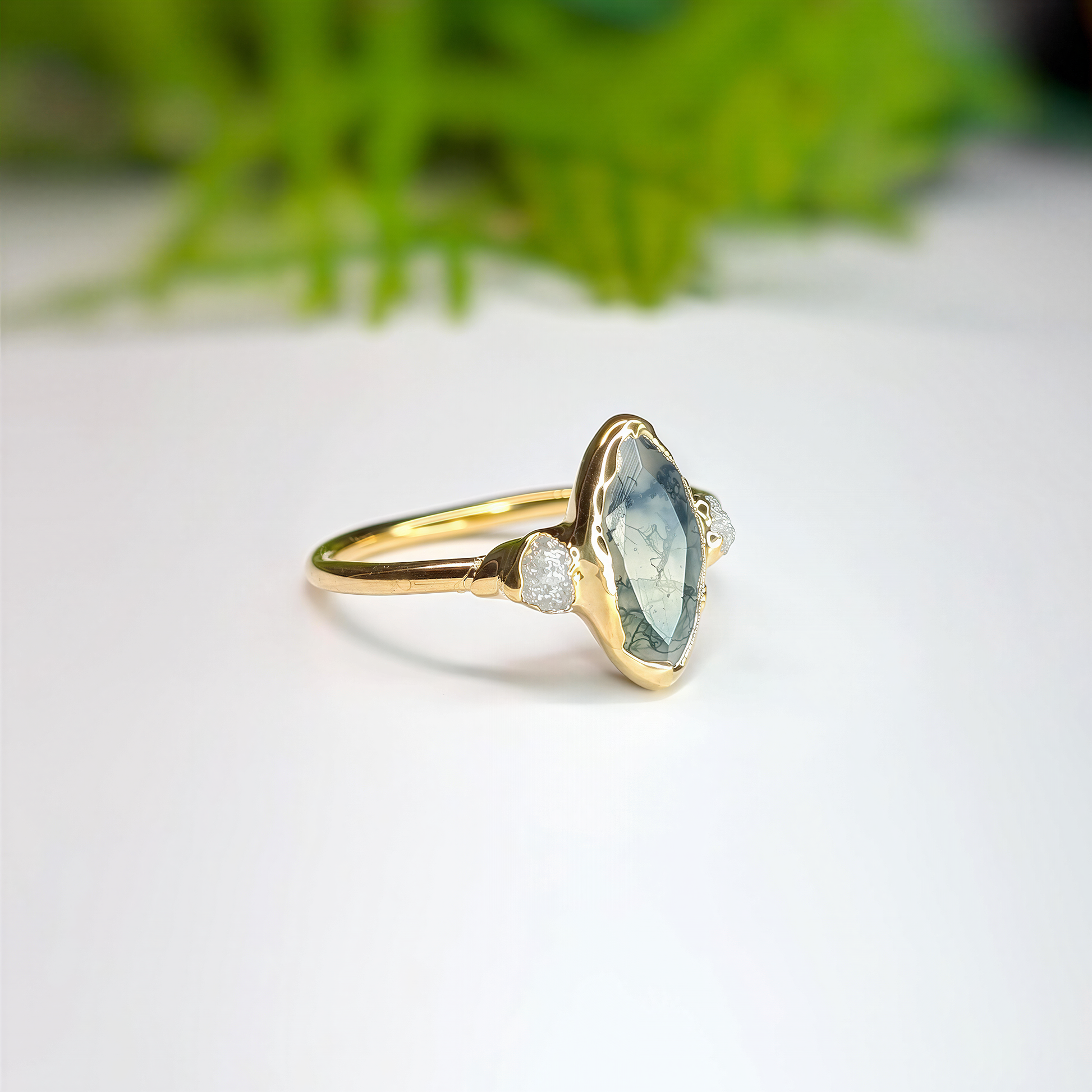 Side view Gold Moss Agate and raw diamond engagement ring in a light background with some greenery