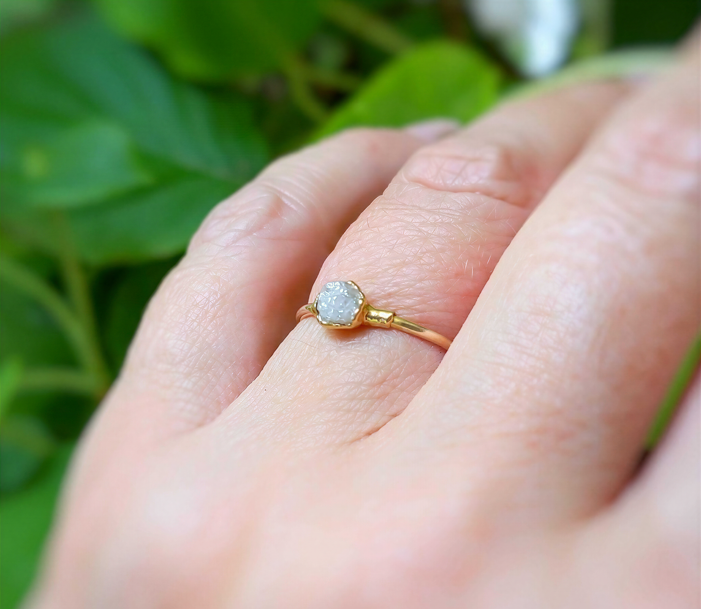 Raw diamond engagement ring on hand, set in solid 14k gold band amidst greenery.