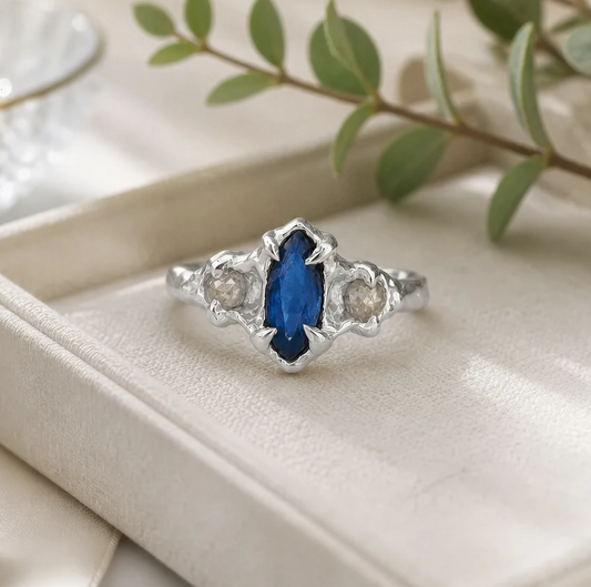 A silver ring with a blue gemstone, placed on a white surface, with a branch of green leaves in the background.