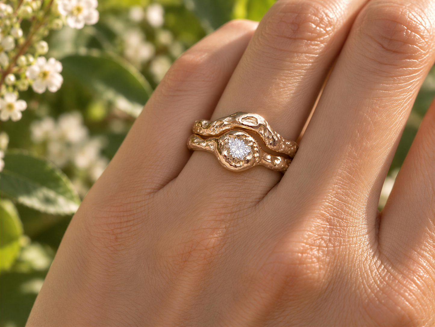 A close-up of a gold ring with a diamond set in the center, worn on a person's hand against a blurred background of greenery.