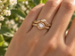 A close-up of a gold ring with a diamond set in the center, worn on a person's hand against a blurred background of greenery.