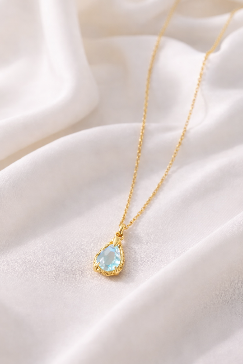 Gold necklace with a teardrop-shaped Aquamarine gemstone pendant on a soft fabric background