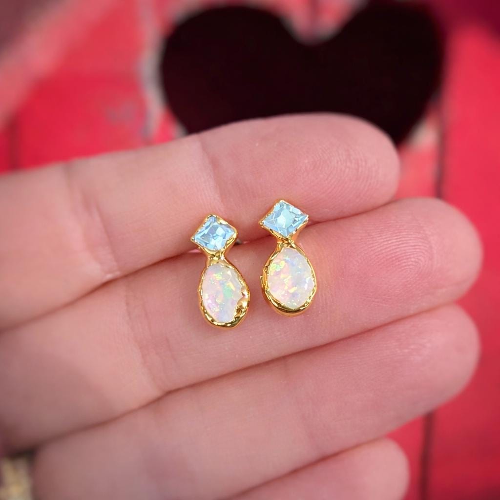 Gold earrings with Aquamarine and Opal gemstones held between fingers against a red background