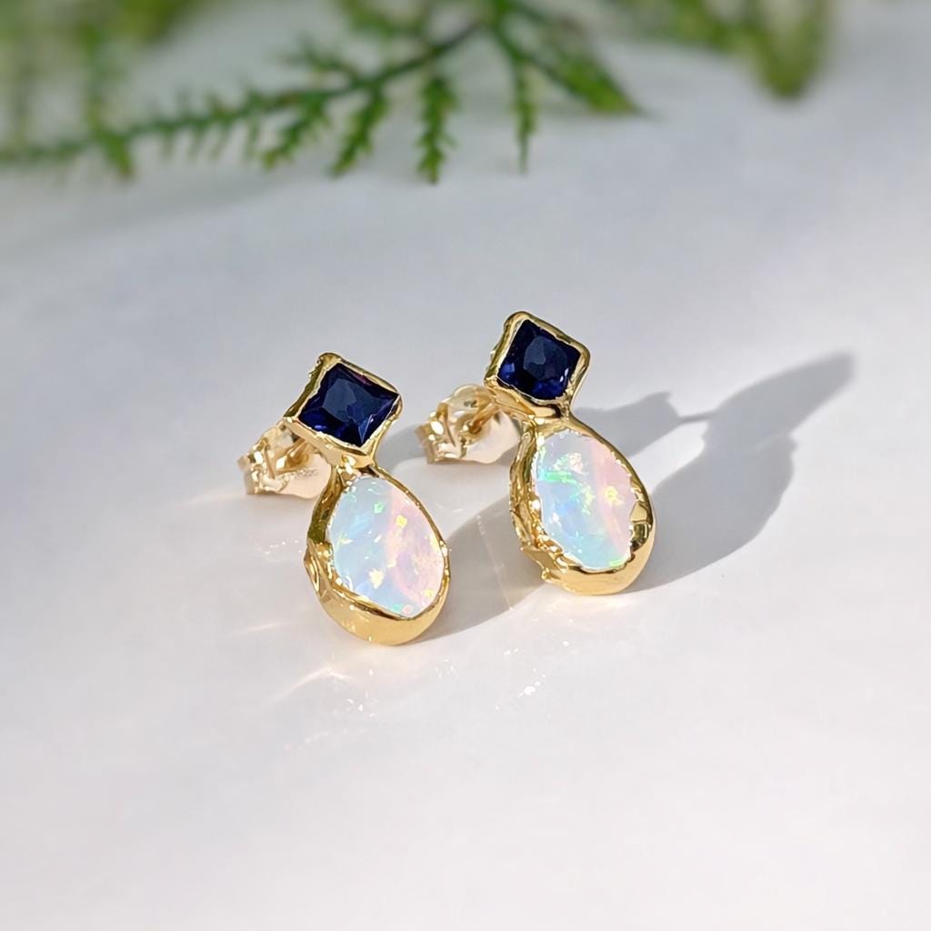 Gold stud earrings with blue Sapphire gemstone and opal teardrop shape on a white background.