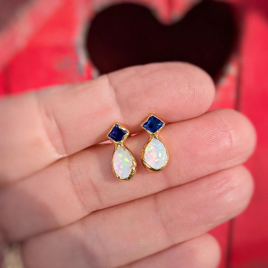 Teardrop-shaped Gold stud earrings with blue Sapphire and Opal held between fingers against a red fabric background.