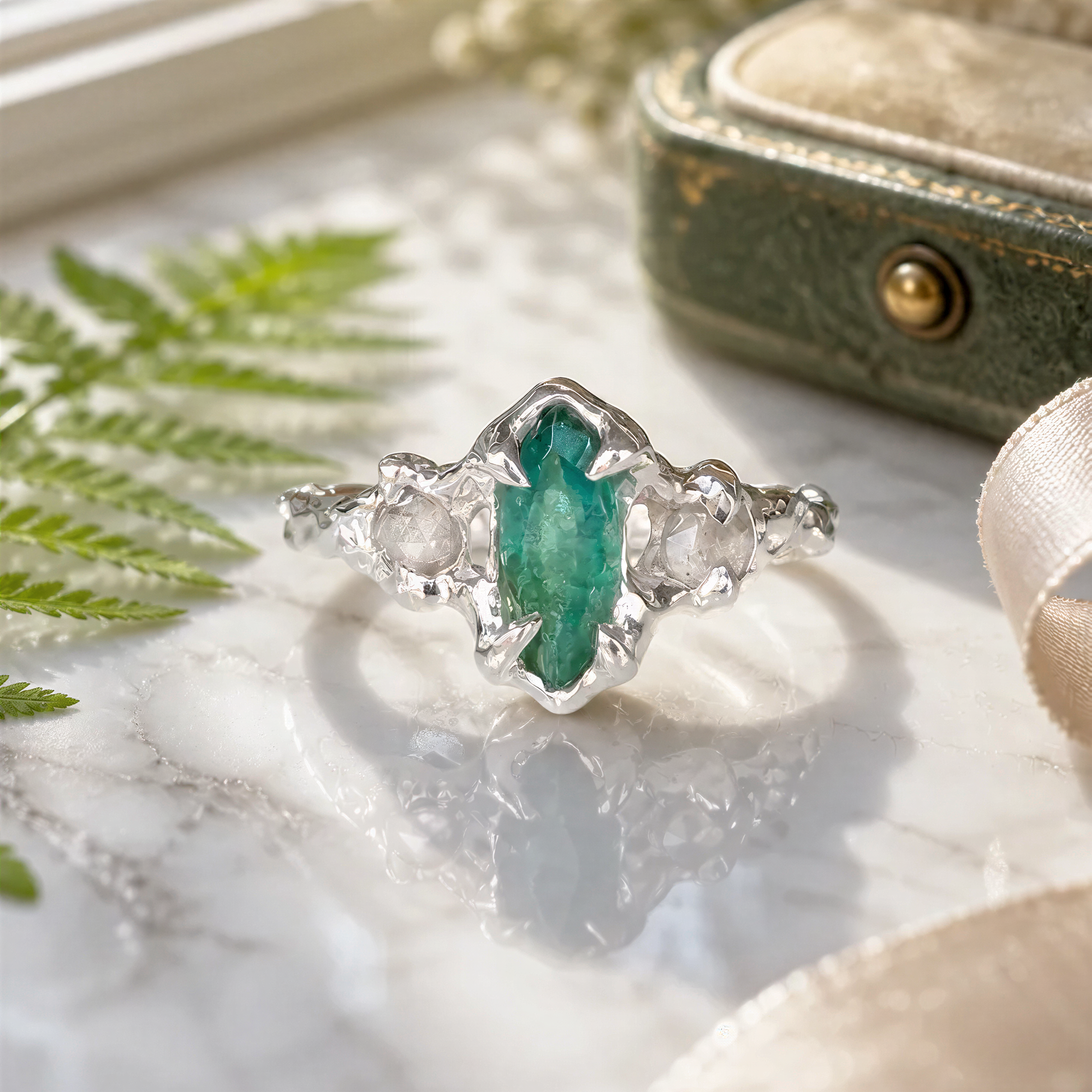A silver ring with a large, vibrant green marquise-shaped Emerald in the center, surrounded by 2 smalle diamonds, sits on a marble surface.