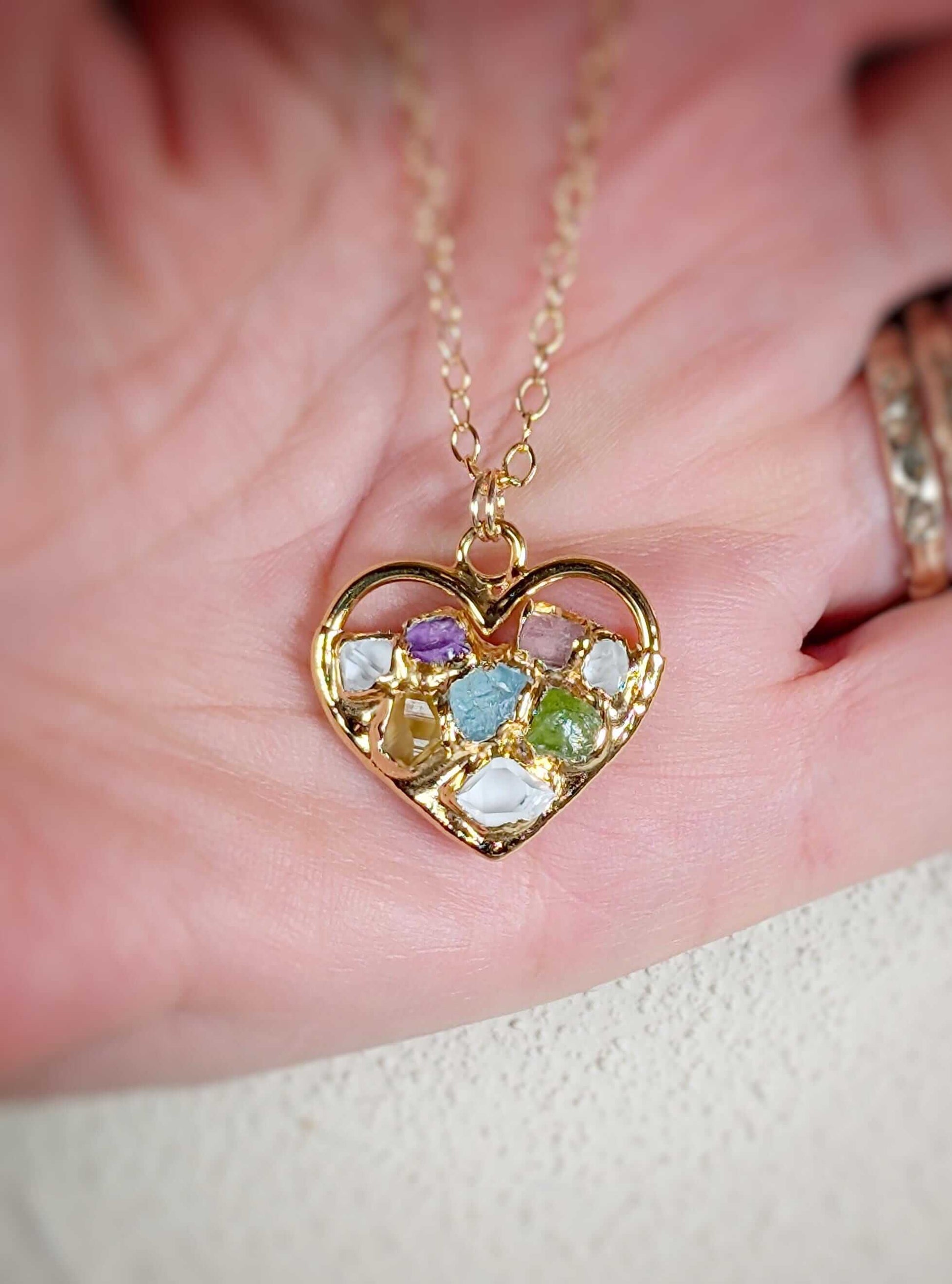 Heart-shaped gold necklace with colorful birthstones representing family members, worn on a hand.