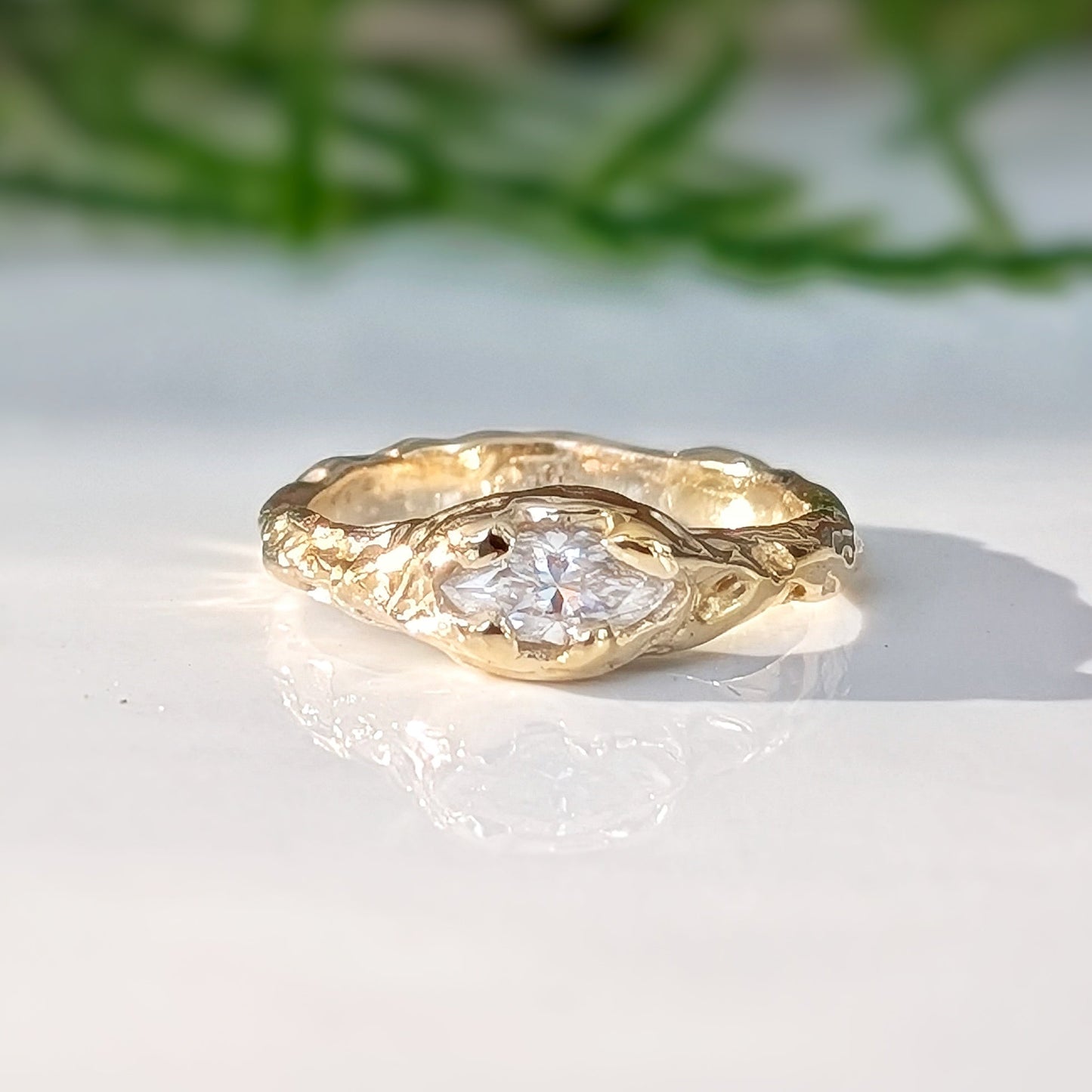 Gold ring with a diamond on a white surface with greenery in the background