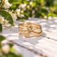 A gold ring with a twisted design sits on a wooden surface, surrounded by white flowers and green foliage.