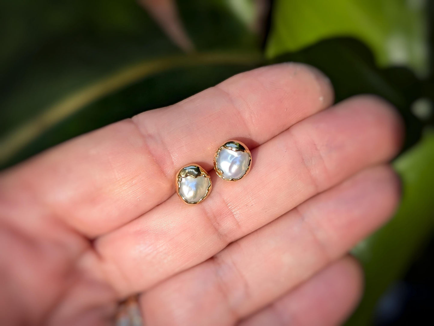 Handcrafted freshwater pearl stud earrings in a unique gold setting, resting on a hand with lush greenery in the background.