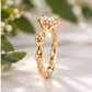 A close-up image of a gold ring with a large central Moissanite, set against a blurred background of green leaves.