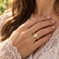 A close-up of a woman's hand wearing a gold ring with a large Moissanite in the center, resting on her heart.