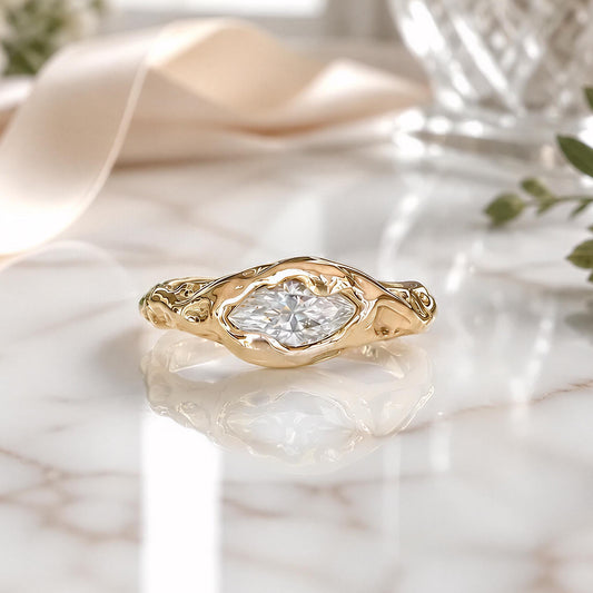A gold ring with a marquise-cut diamond set in an organic, textured bezel, resting on a white marble surface with a soft beige ribbon and blurred decorative elements in the background.