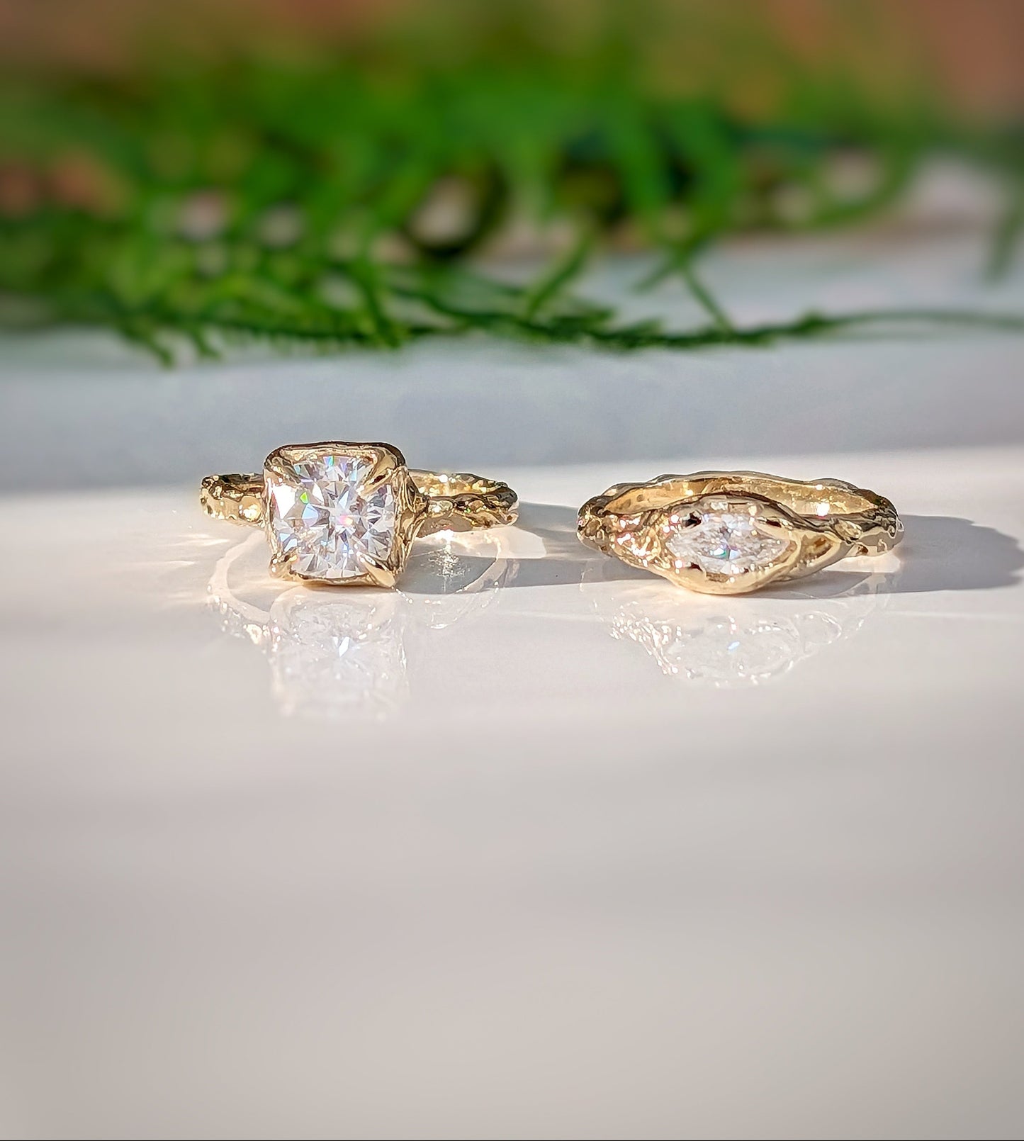 Two gold Moissanite rings on a light surface with greenery in the background