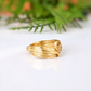 A gold ring with a leaf-like design sits on a white surface, with a blurred green plant in the background.