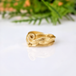A gold ring with a swirling textured design sits on a white surface, with a blurred green plant in the background.