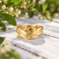 A gold ring with a textured, curled design sits on a wooden surface, surrounded by green foliage.