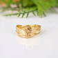 A gold ring with a swirling design sits on a white surface, with a blurred green plant in the background.