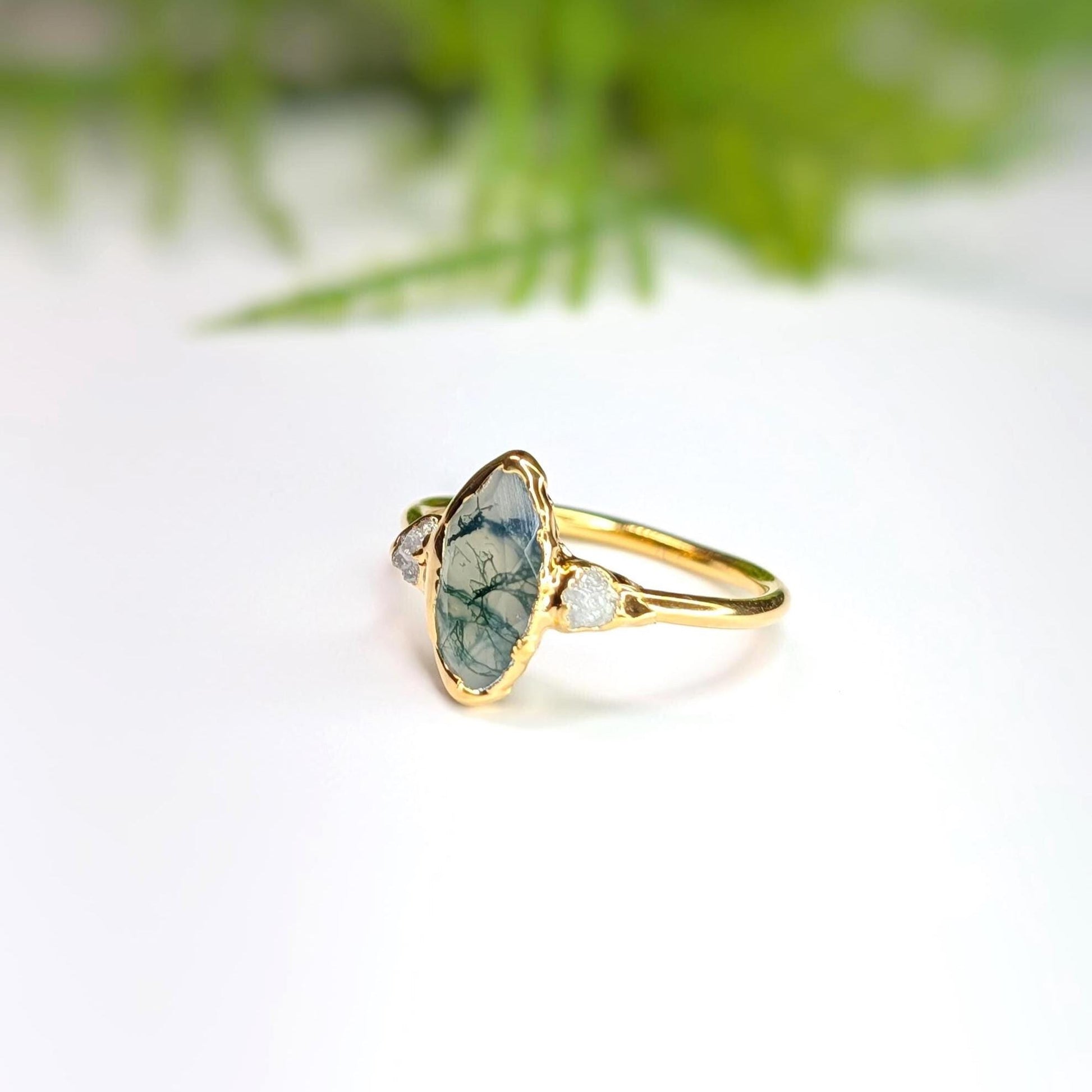 Side view of a Gold Moss Agate and raw diamond engagement ring in a light background with some greenery